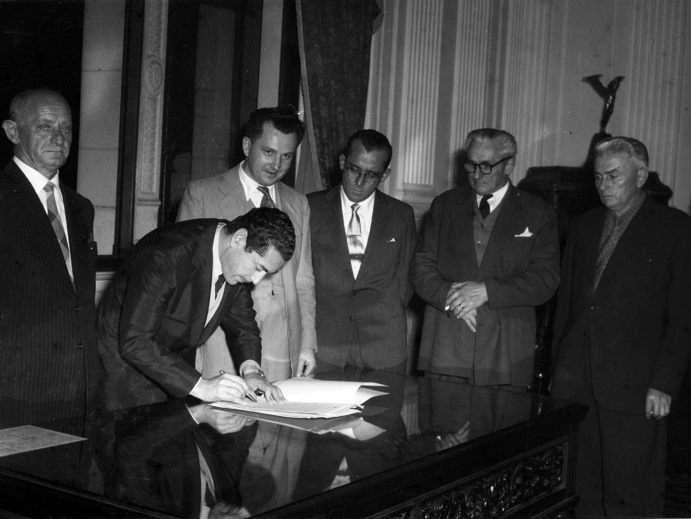 Assinatura da lei de criação do município de Dois Irmãos, em Porto Alegre, em 1959: um homem assina o documento sobre a mesa enquanto cinco integrantes da comissão emancipacionista acompanham em pé
